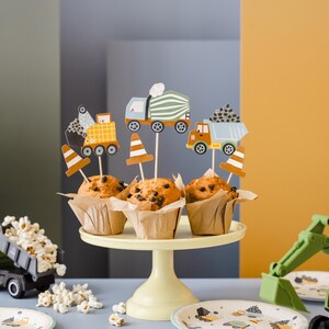 May include: A display of muffins with construction-themed toppers, including trucks and traffic cones. The muffins sit on a yellow cake stand, with a toy truck and excavator nearby. The scene is set against a backdrop of yellow, gray, and blue.