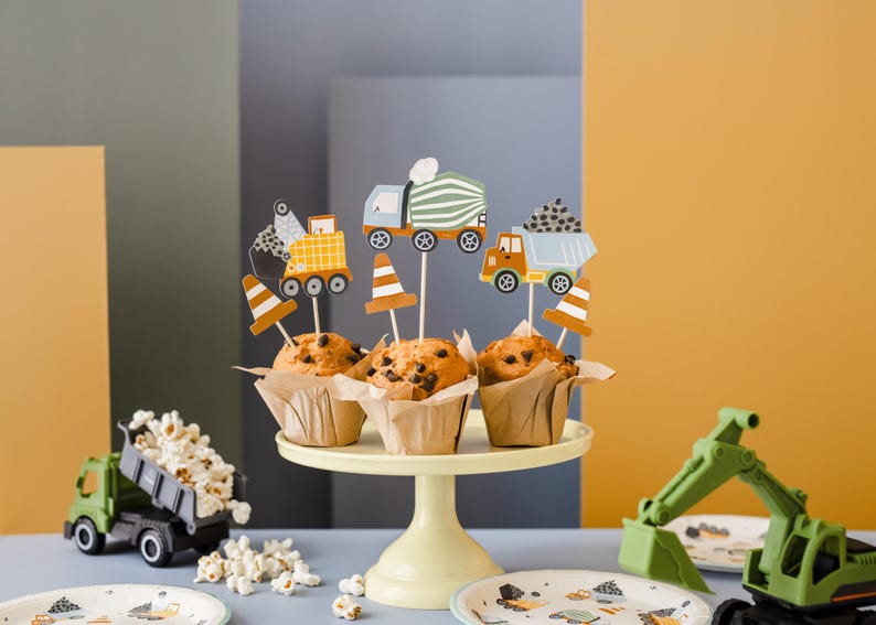 May include: A display of muffins with construction-themed toppers, including trucks and traffic cones. The muffins sit on a yellow cake stand, with a toy truck and excavator nearby. The scene is set against a backdrop of yellow, gray, and blue.