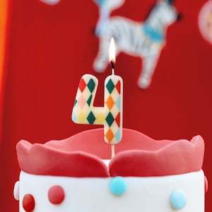 Könnte beinhalten: Eine bunte Zahl vier Kerze mit einem Diamantenmuster. Die Kerze ist angezündet und sitzt auf einem rosa und weißen Kuchen.