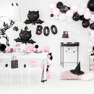 K&ouml;nnte beinhalten: Eine Halloween-Party-Tischdekoration in Schwarz und Rosa. Es gibt schwarze und wei&szlig;e Ballons, einen schwarzen und wei&szlig;en Skelettballon, schwarze Fledermausballons und einen schwarzen Hexenhut. Der Tisch ist mit schwarzen und wei&szlig;en Tellern, Servietten und Bechern gedeckt. Es gibt auch schwarze und wei&szlig;e Geschenkt&uuml;ten mit einem Totenkopfdesign.