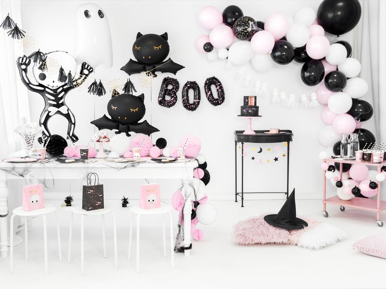 K&ouml;nnte beinhalten: Eine Halloween-Party-Tischdekoration in Schwarz und Rosa. Es gibt schwarze und wei&szlig;e Ballons, einen schwarzen und wei&szlig;en Skelettballon, schwarze Fledermausballons und einen schwarzen Hexenhut. Der Tisch ist mit schwarzen und wei&szlig;en Tellern, Servietten und Bechern gedeckt. Es gibt auch schwarze und wei&szlig;e Geschenkt&uuml;ten mit einem Totenkopfdesign.