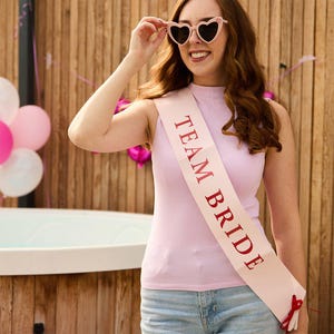 May include: A light pink sleeveless top and light blue denim shorts. A pink sash with the text "TEAM BRIDE" in red is draped across the front. The person is wearing heart-shaped sunglasses.