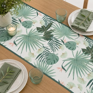 May include: A rectangular table runner featuring a white background with a repeating botanical pattern of green palm leaves, monstera leaves, and small pink accents. The runner has a dark green border and is laid out on a wooden surface.