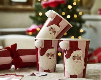 May include: A stack of red and pink striped paper cups with Santa Claus faces and white pom-poms. The cups are on a table with Christmas decorations, including a red cracker and Santa Claus cutouts.