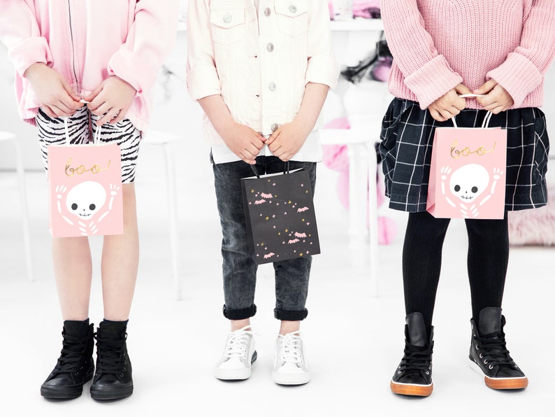 May include: Three children holding pink and black Halloween treat bags with a skeleton and bat design. The bags say "Boo!" on them.
