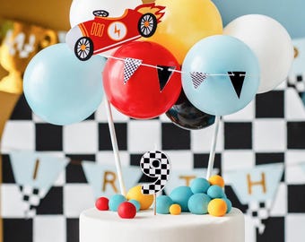 May include: A decorated cake with a racing theme. It features a red race car topper, balloons in white, yellow, red, and blue, and a number 9 candle. The cake is decorated with blue and yellow candies and a checkered flag banner.