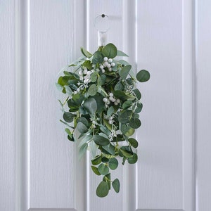 Op de afbeelding: Een hangende eucalyptus- en bessenslinger. De slinger heeft donkergroene eucalyptusbladeren en kleine witte bessen. Het is bevestigd aan een metalen ring met een wit lint om op te hangen. De slinger wordt getoond tegen een witte deur met panelen.