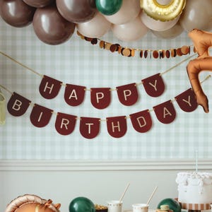 Può includere: Decorazione per festa di compleanno con uno striscione bordeaux "HAPPY BIRTHDAY". Palloncini marroni, verdi e dorati sono sospesi sopra. Una torta bianca con una candela è su un tavolo.