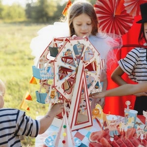 May include: A colorful wooden Ferris wheel toy with red, white, and blue accents. The toy is surrounded by children dressed in festive attire, including a girl wearing a white dress and a boy wearing a striped shirt. The scene is set against a backdrop of red and white striped fabric and a green lawn.