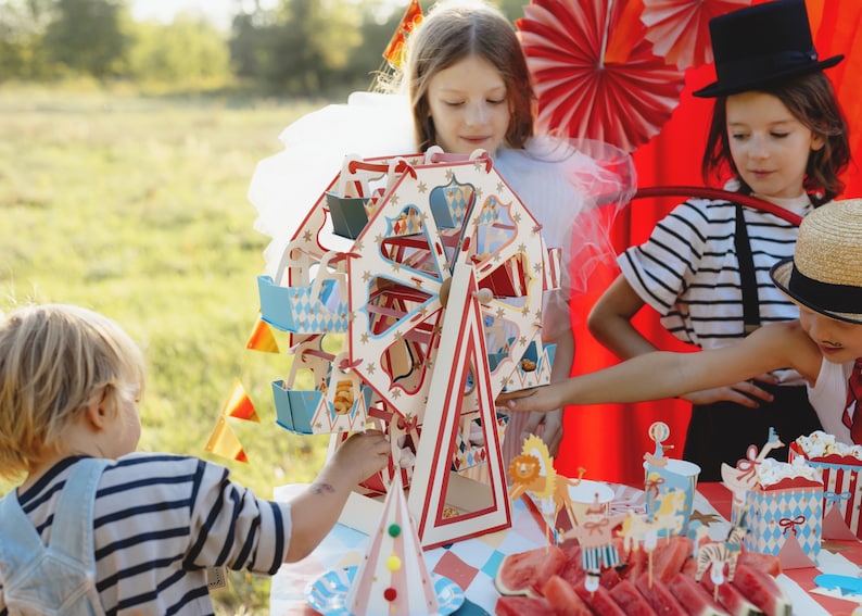 May include: A colorful wooden Ferris wheel toy with red, white, and blue accents. The toy is surrounded by children dressed in festive attire, including a girl wearing a white dress and a boy wearing a striped shirt. The scene is set against a backdrop of red and white striped fabric and a green lawn.