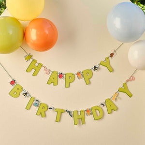 May include: A green and yellow banner with the words "Happy Birthday" decorated with small insect cut-outs. The banner is hanging on a white wall with balloons in the background.