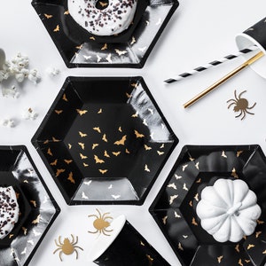 May include: Black hexagonal plates and cups with gold bat patterns are arranged on a white surface. The plates hold donuts and a white pumpkin. Gold and black straws and spider cutouts complete the Halloween party theme.