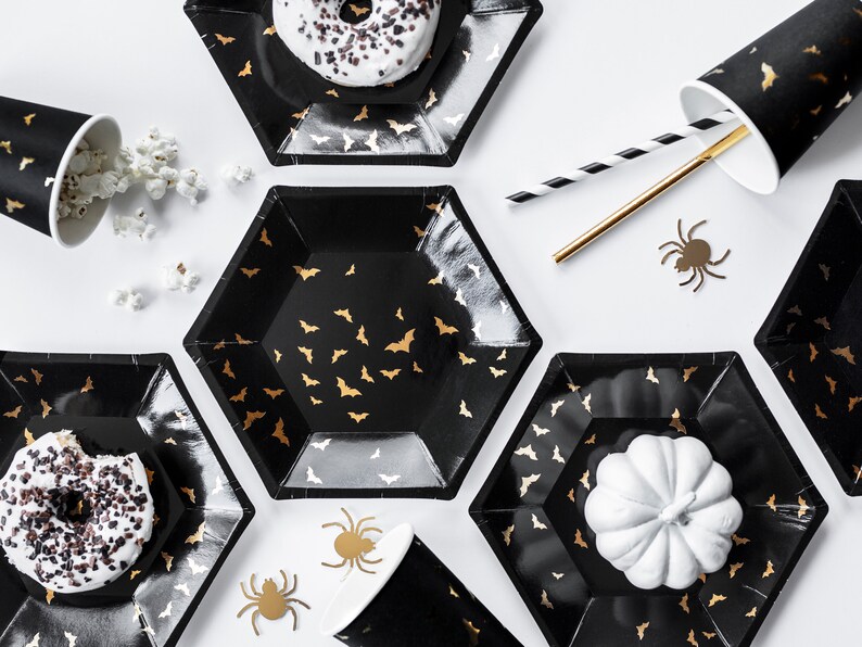 May include: Black hexagonal plates and cups with gold bat patterns are arranged on a white surface. The plates hold donuts and a white pumpkin. Gold and black straws and spider cutouts complete the Halloween party theme.