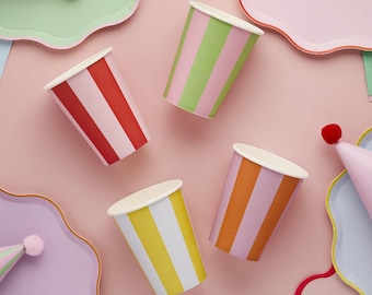 May include: Overhead shot of colorful party supplies on a pink background. Five paper cups with vertical stripes in red, green, orange, and yellow are scattered around. Party hats and plates are also visible.