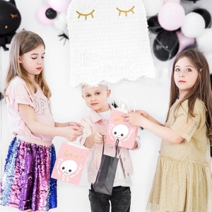 May include: Three children are holding pink and black Halloween treat bags with a skeleton design and the word "Boo!" printed on them. The bags are decorated with gold glitter. The children are standing in front of a white wall with black and white balloons.