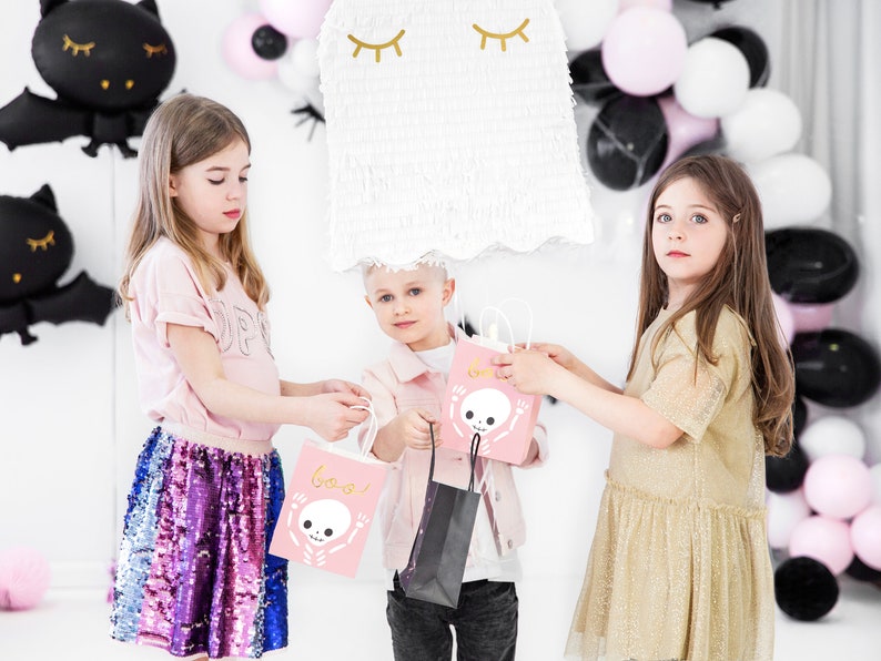 May include: Three children are holding pink and black Halloween treat bags with a skeleton design and the word "Boo!" printed on them. The bags are decorated with gold glitter. The children are standing in front of a white wall with black and white balloons.