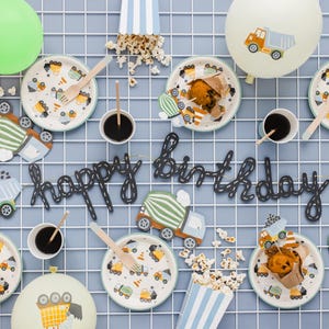 May include: A festive birthday party setup with construction-themed decorations. Includes plates, cups, wooden utensils, popcorn, and balloons. The phrase "happy birthday" is spelled out in black script. Plates feature construction vehicle designs.