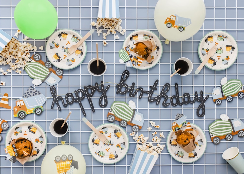 May include: A festive birthday party setup with construction-themed decorations. Includes plates, cups, wooden utensils, popcorn, and balloons. The phrase "happy birthday" is spelled out in black script. Plates feature construction vehicle designs.