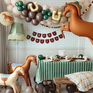 以下が含まれることがあります： 馬をテーマにした誕生日のパーティーシーン。茶色の大きな馬の風船、緑と白のストライプのテーブルクロスをかけたテーブル、そして「HAPPY BIRTHDAY」と書かれたバナーが含まれています。