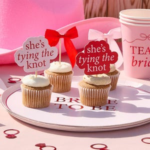 Peut inclure: Des cupcakes avec glaçage blanc et des décorations de nœuds rouges, chacun surmonté d'un pic avec l'inscription "she's tying the knot". Les cupcakes sont sur une assiette rose avec le texte "BRIDE TO BE", à côté d'un gobelet rose.