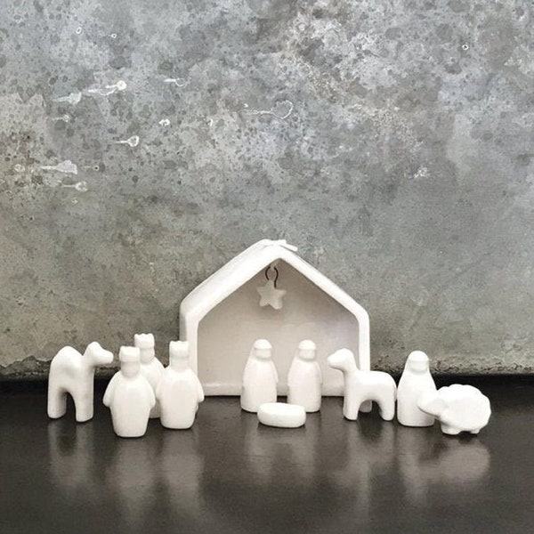 May include: A white ceramic nativity set featuring a stable with a hanging star, and figurines of the Holy Family, wise men, animals, and a manger. The set is displayed on a dark surface against a gray background.