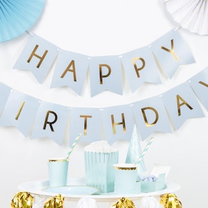 Könnte beinhalten: Eine Geburtstagsfeier mit hellblauen Dekorationen. Ein Banner mit der Aufschrift "HAPPY BIRTHDAY" in goldenen Buchstaben ist oben angebracht. Partyzubehör wie Becher, Teller und eine Popcornbox sind hellblau mit goldenen Akzenten. Papierfächer und andere Dekorationen ergänzen die Szene.