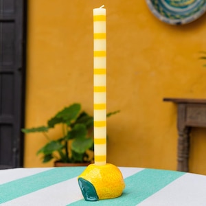 May include: A decorative candle featuring yellow and white stripes, placed on a lemon-shaped holder. The holder is yellow with a green accent. The background includes a yellow wall and a striped tablecloth.