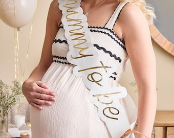 May include: A white sash with gold lettering that reads "Mummy to be" is worn over a cream-colored dress. The sash has a scalloped edge and a brown ribbon tie. A cream-colored balloon is visible in the background.