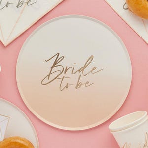 May include: A round, ombre-colored plate with the words "Bride to be" in gold script. The plate has a gradient effect, transitioning from white to a peach color. Other party supplies are visible in the background.