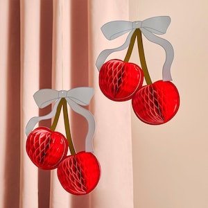 May include: Two hanging decorations shaped like pairs of cherries. Each cherry features a bright red honeycomb paper structure, brown stems, and a light gray paper bow at the top. The decorations are suspended by thin strings for display.