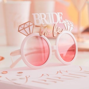 May include: White plastic sunglasses with pink tinted lenses and a gold glitter "Bride to Be" sign with diamond accents.