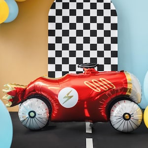 以下が含まれることがあります： 白い車輪と金色のアクセントが特徴の、レーシングカーの形をした赤いホイルバルーン。車には稲妻のエンブレムと金色のスピードラインがあります。背景には、白黒の市松模様と風船があります。