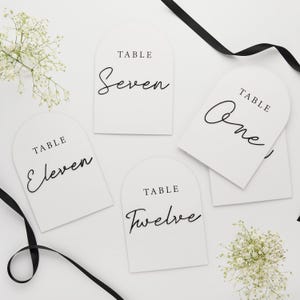 May include: White arched table number cards with black script. The cards are labeled with the numbers "One", "Seven", "Eleven", and "Twelve". Black ribbon and white flowers are arranged around the cards.