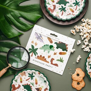 May include: A still-life image featuring dinosaur-themed party supplies. Includes temporary tattoos, scalloped-edge plates, and popcorn. The plates and tattoos feature dinosaur and palm tree illustrations. The words "DINOSAUR PARTY" are visible on the tattoo packaging.