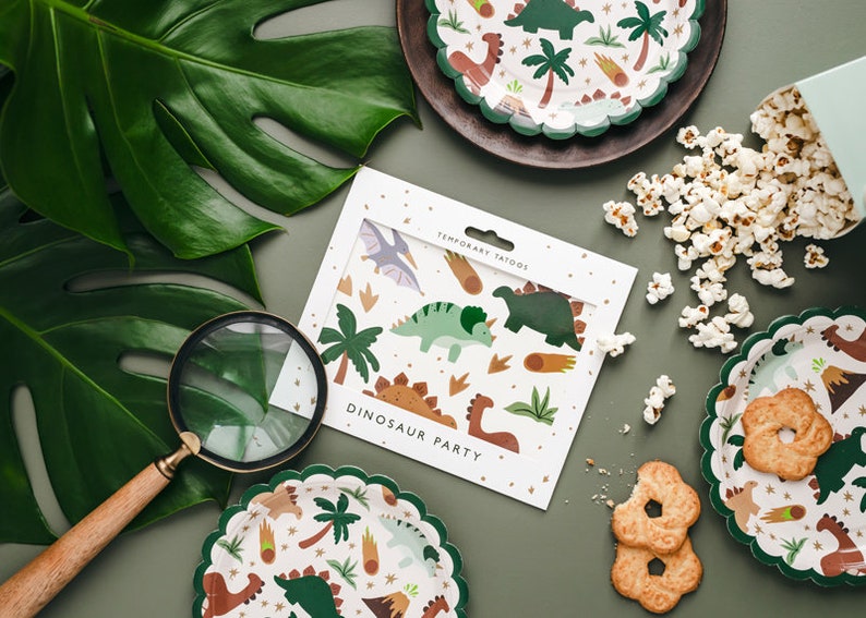 May include: A still-life image featuring dinosaur-themed party supplies. Includes temporary tattoos, scalloped-edge plates, and popcorn. The plates and tattoos feature dinosaur and palm tree illustrations. The words "DINOSAUR PARTY" are visible on the tattoo packaging.