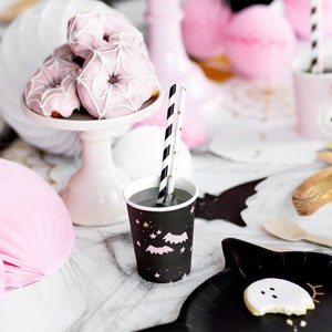 May include: A festive Halloween party scene with pink spiderweb donuts on a white cake stand, black cups with bats and stars, and black and white striped straws. Pink honeycomb decorations and a black bat-shaped plate with a ghost cookie complete the spooky theme.