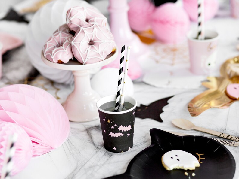 May include: A festive Halloween party scene with pink spiderweb donuts on a white cake stand, black cups with bats and stars, and black and white striped straws. Pink honeycomb decorations and a black bat-shaped plate with a ghost cookie complete the spooky theme.