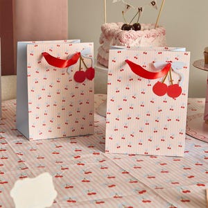 May include: Two rectangular paper gift bags featuring a light pink and white vertical striped pattern with small red cherry illustrations. Each bag has a bright red satin ribbon handle and a decorative paper cherry cutout with a white bow attached.