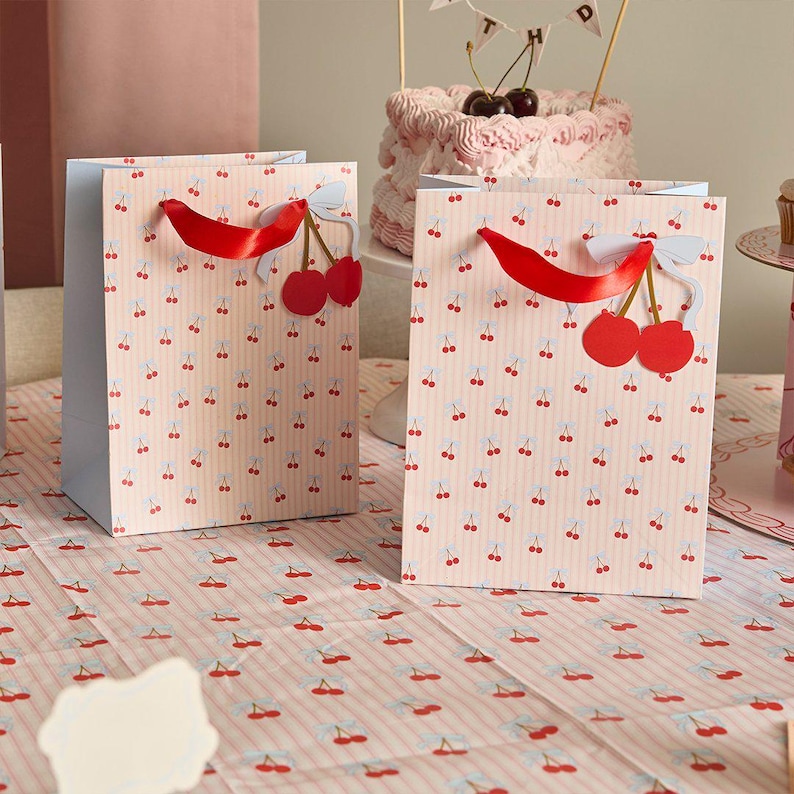 May include: Two rectangular paper gift bags featuring a light pink and white vertical striped pattern with small red cherry illustrations. Each bag has a bright red satin ribbon handle and a decorative paper cherry cutout with a white bow attached.