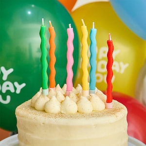 May include: A frosted cake decorated with a selection of colourful, wavy birthday candles. The candles are green, orange, pink, yellow, blue, and red. Balloons in the background enhance the celebratory theme.