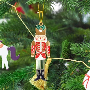 May include: A festive Christmas tree decoration depicting a nutcracker soldier. The ornament features a red coat, white details, and a gold crown, with black boots. It hangs from a gold string and tassel, set against a backdrop of green pine needles.