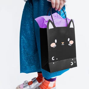 May include: A child wearing blue sparkly pants and silver sandals holds a black paper bag with a black cat face and white stars. The bag has a purple tissue paper inside.