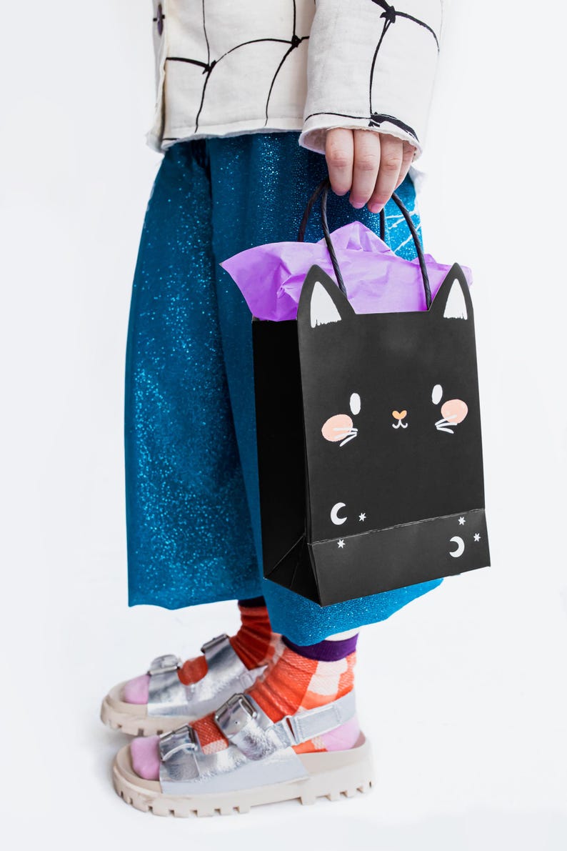 May include: A child wearing blue sparkly pants and silver sandals holds a black paper bag with a black cat face and white stars. The bag has a purple tissue paper inside.