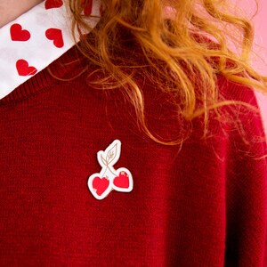 Könnte beinhalten: Ein roter Pullover mit einem weißen Hemd mit Kragen darunter. Das Hemd hat ein Muster aus roten Herzen. Eine kirschförmige Brosche ist am Pullover befestigt.