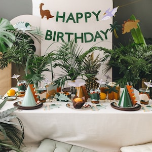 May include: A festive birthday party table setting with a dinosaur theme. The table is set with plates, cups, and party hats. A banner reads "HAPPY BIRTHDAY" with dinosaur cutouts. Tropical plants and a pineapple add to the decor.
