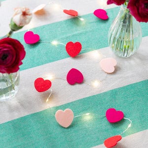 May include: A string of felt heart decorations in shades of red, pink, and light pink, interspersed with tiny fairy lights. The hearts are arranged on a striped green and white tablecloth, with small vases of flowers in the background.