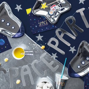 May include: A silver space party banner with the words "SPACE PARTY" in large letters. The banner is decorated with silver stars and is hanging on a blue background. There are also silver rocket-shaped plates, a silver star-shaped plate, and a silver cup with the word "SPACE" on it.