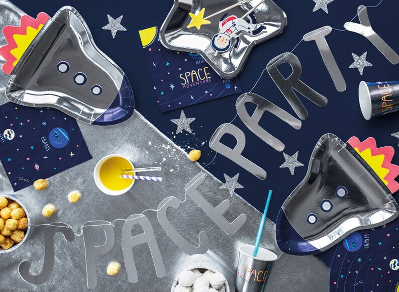 May include: A silver space party banner with the words "SPACE PARTY" in large letters. The banner is decorated with silver stars and is hanging on a blue background. There are also silver rocket-shaped plates, a silver star-shaped plate, and a silver cup with the word "SPACE" on it.