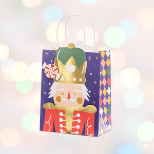 Könnte beinhalten: Eine violette Geschenktüte mit einem Nussknacker-Motiv. Der Nussknacker trägt eine goldene Krone und eine rot-goldene Uniform. Die Tasche hat einen weißen Henkel und einen Lebkuchenmann-Anhänger.