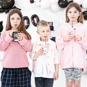 May include: Three children at a Halloween party, each holding a cup with a straw. The children are wearing pink clothing. The background includes black and white balloons and Halloween decorations. One child wears a pink sweater, another a pink jacket, and the third a pink zip-up hoodie.